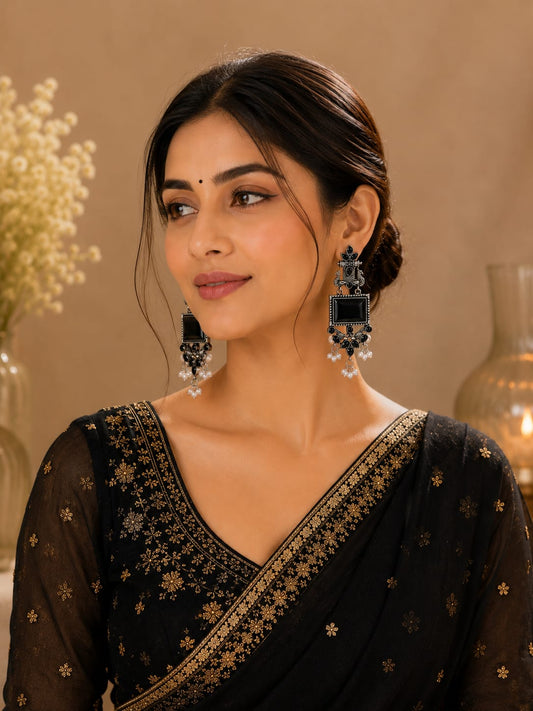 Model in a black saree with gold zari wearing long oxidised peacock earrings with black stones and pearl drops