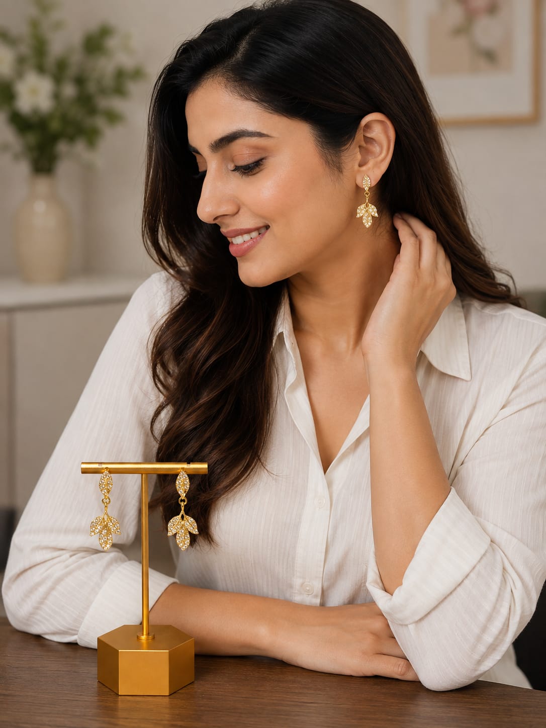 Model in a white shirt wearing gold-plated crystal leaf drop earrings beside a gold display stand