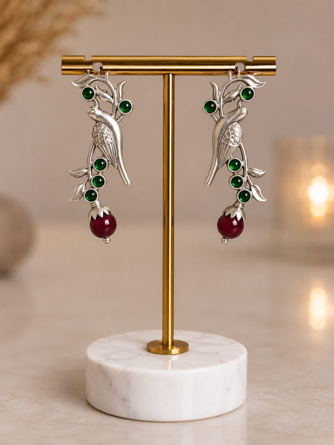 Oxidised silver parrot drop earrings with green glass stones and a maroon bead drop on a brass display stand