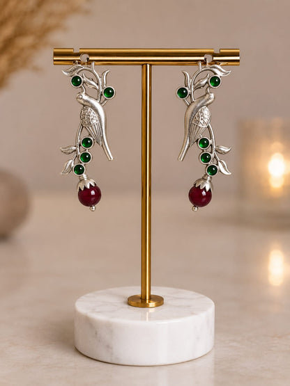 Oxidised silver parrot drop earrings with green glass stones and a maroon bead drop on a brass display stand
