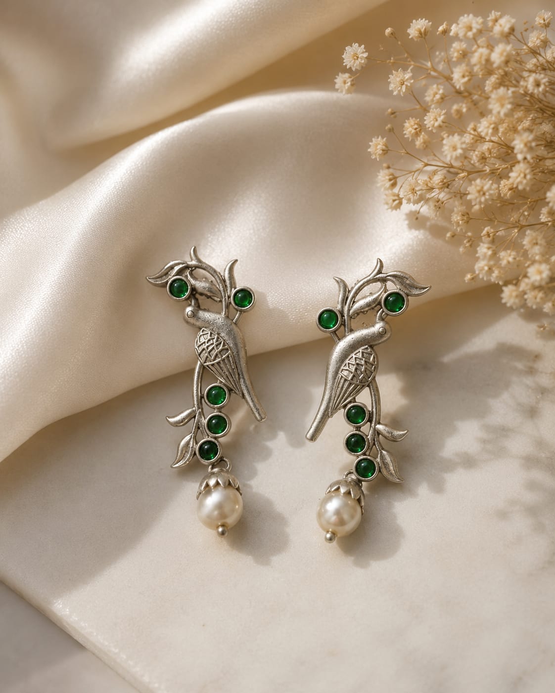 Oxidised silver parrot drop earrings with green glass stones and a white pearl drop on ivory satin