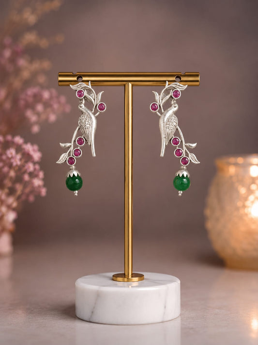 Oxidised silver parrot drop earrings with ruby pink stones and a green bead drop on a brass display stand