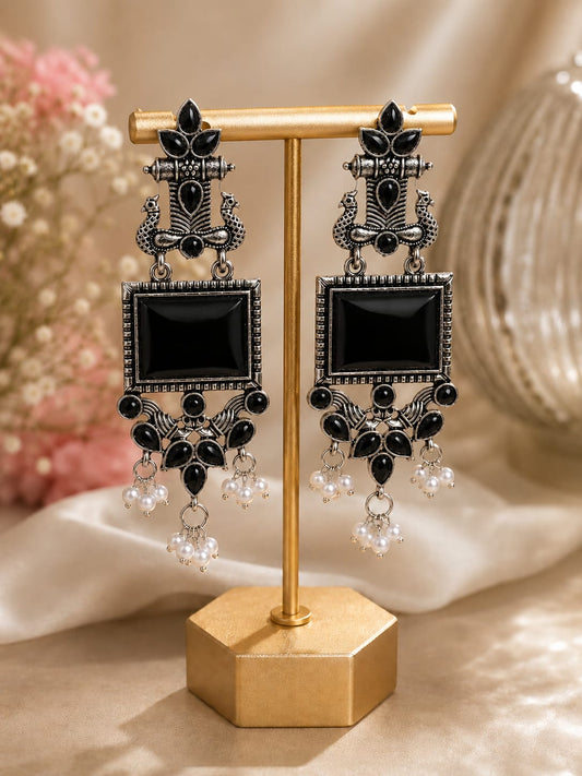 Oxidised silver peacock chandelier earrings with black stones and faux pearl clusters on a gold display stand