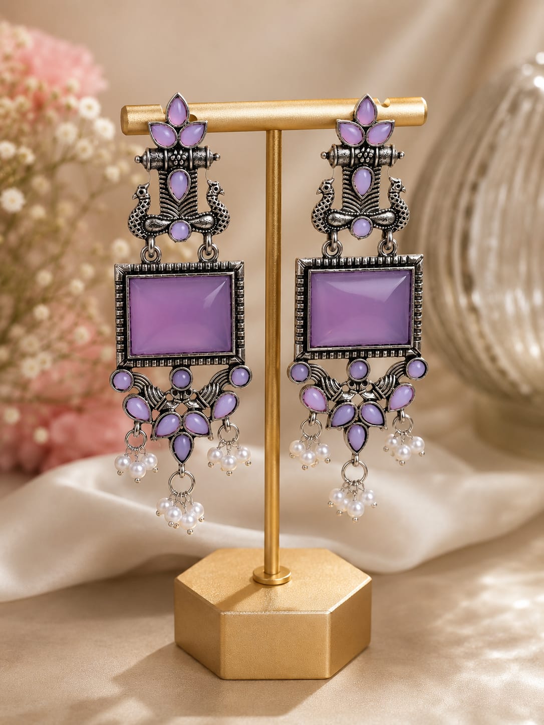 Oxidised silver peacock chandelier earrings with lilac stones and faux pearl clusters on a gold display stand