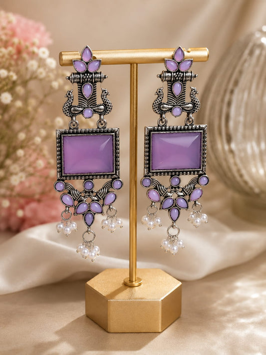 Oxidised silver peacock chandelier earrings with lilac stones and faux pearl clusters on a gold display stand