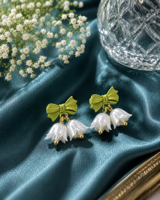 Green bow and pearl lily bell earrings on teal satin beside a cut-glass vase